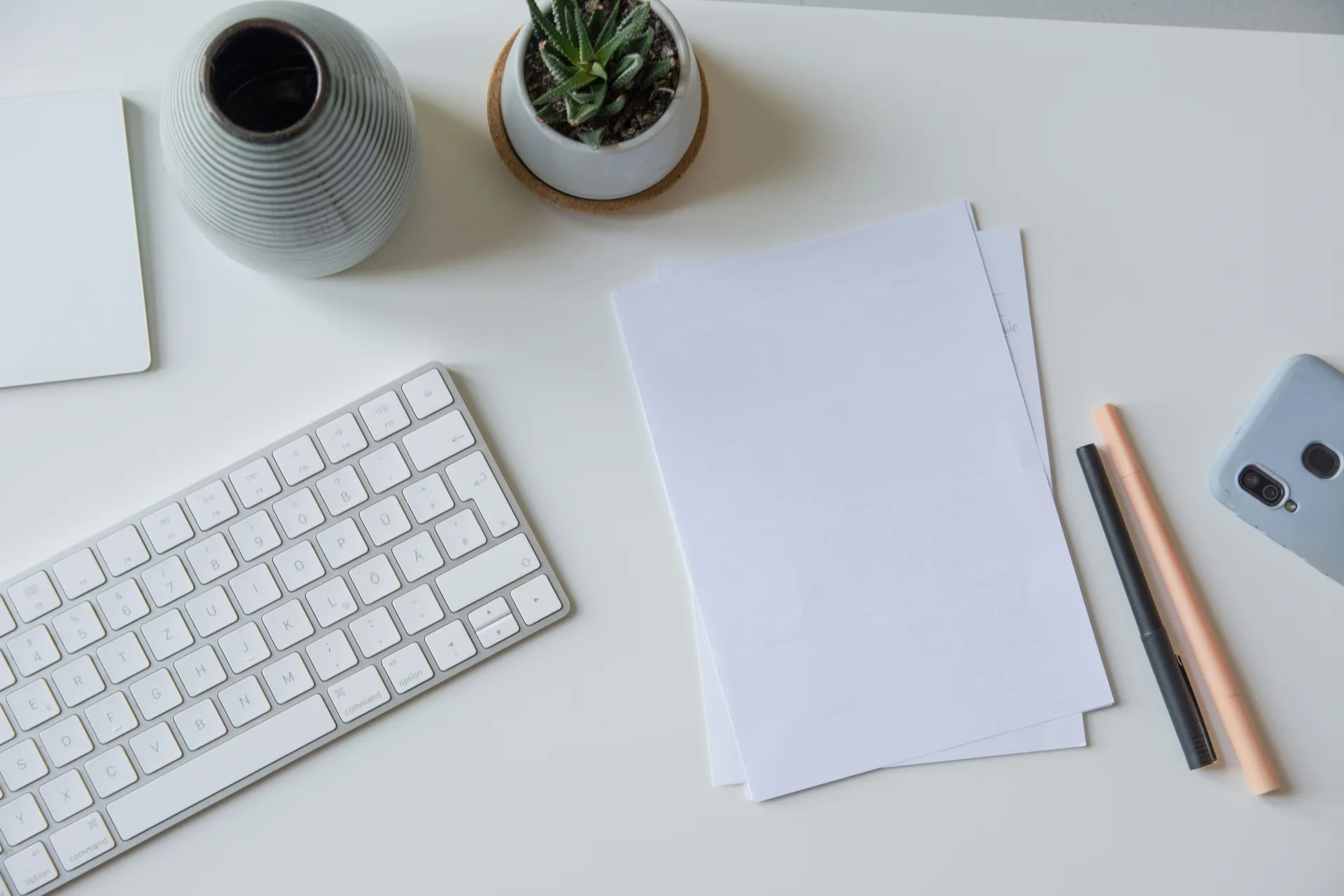 An overhead view of a bright back-office desk with paperwork, a keyboard, pens, and a small plant